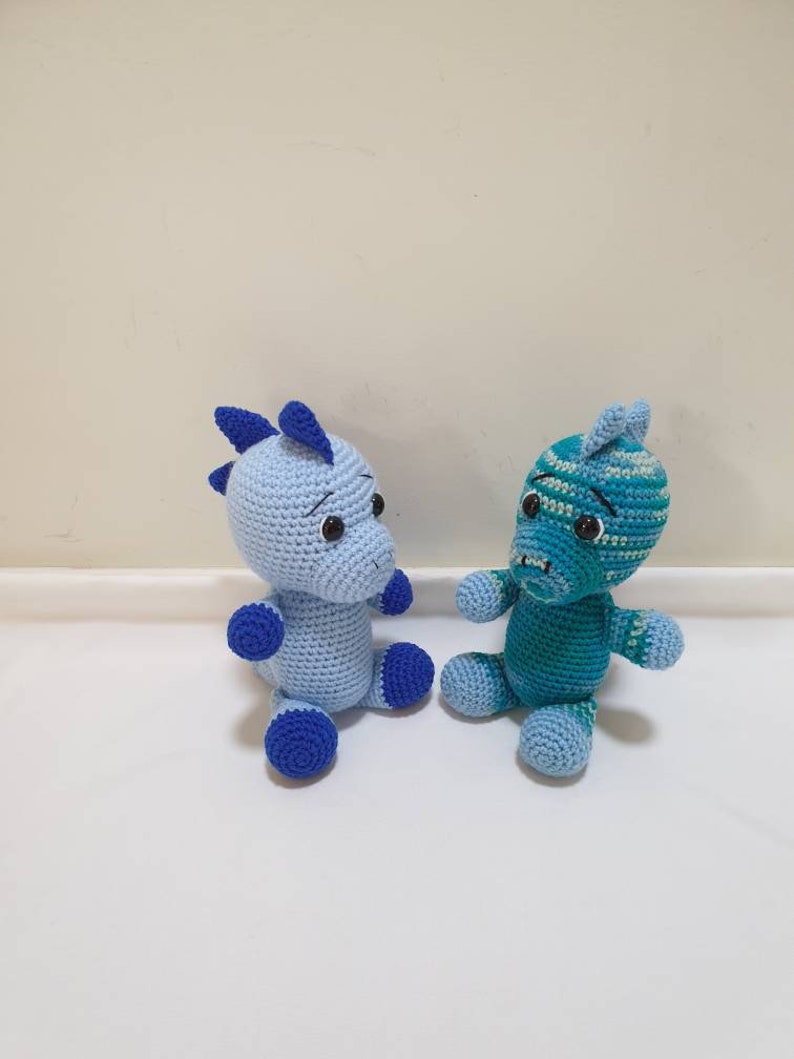 May include: Two crocheted stuffed animals, one blue and one teal, sitting on a white surface. Both animals have blue eyes and a blue nose.