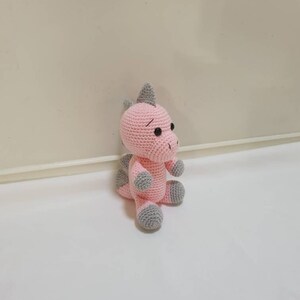 May include: A pink crocheted dinosaur toy with grey accents. The dinosaur has a small grey horn on its head and grey feet.