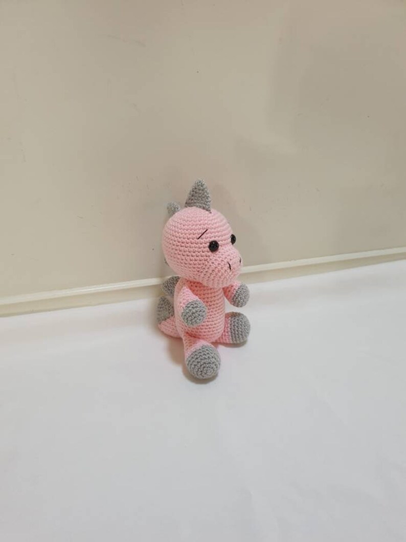 May include: A pink crocheted dinosaur toy with grey accents. The dinosaur has a small grey horn on its head and grey feet.