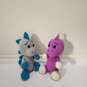May include: Two crocheted dinosaur toys, one gray with teal accents and one purple with white accents. Both toys have black eyes and are sitting on a white surface.