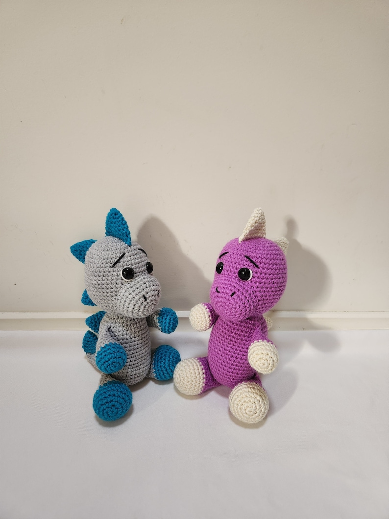 May include: Two crocheted dinosaur toys, one gray with teal accents and one purple with white accents. Both toys have black eyes and are sitting on a white surface.