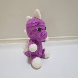 May include: A purple crocheted dinosaur toy with white accents on the ears, feet, and tail. The dinosaur has black eyes and a stitched-on smile.