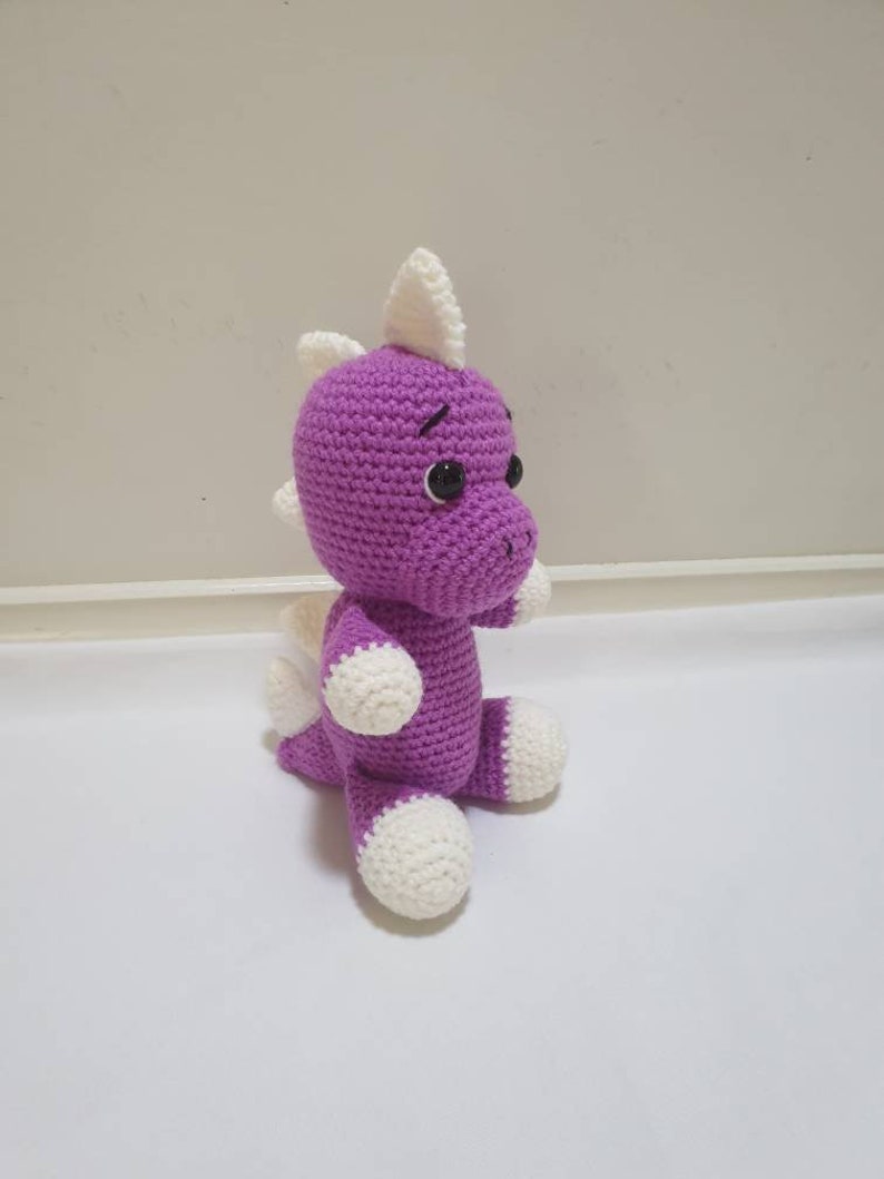 May include: A purple crocheted dinosaur toy with white accents on the ears, feet, and tail. The dinosaur has black eyes and a stitched-on smile.