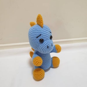 May include: A blue crocheted dinosaur toy with yellow accents. The dinosaur has a small, pointed nose and large, black eyes.