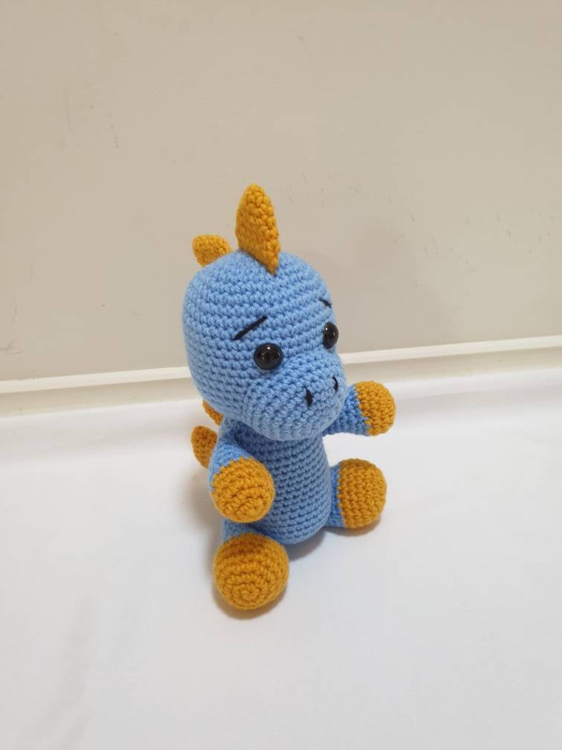 May include: A blue crocheted dinosaur toy with yellow accents. The dinosaur has a small, pointed nose and large, black eyes.