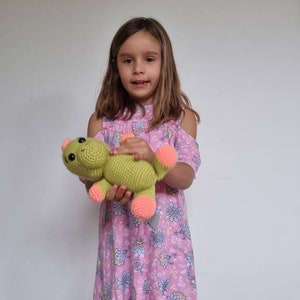 May include: A young girl in a pink floral dress holds a green crocheted stuffed animal with pink accents. The stuffed animal has a friendly face and is shaped like a frog.