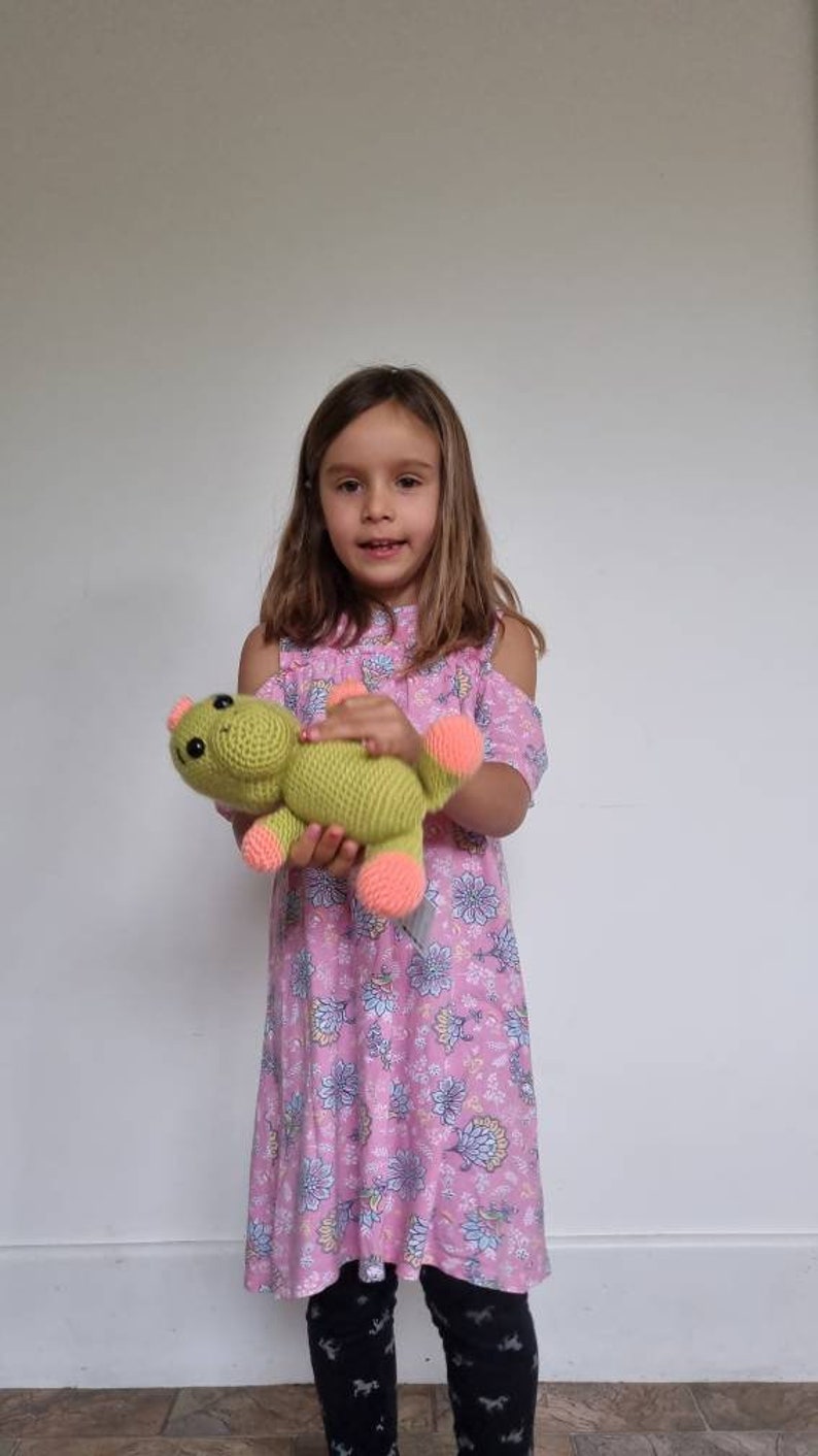 May include: A young girl in a pink floral dress holds a green crocheted stuffed animal with pink accents. The stuffed animal has a friendly face and is shaped like a frog.