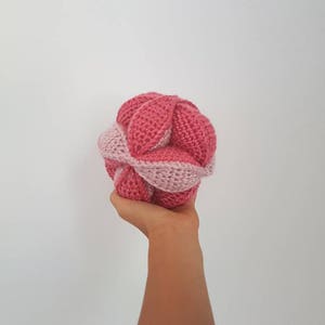 May include: A pink and light pink crocheted ball with a unique, intricate design. The ball is made of yarn and has a soft, textured surface.