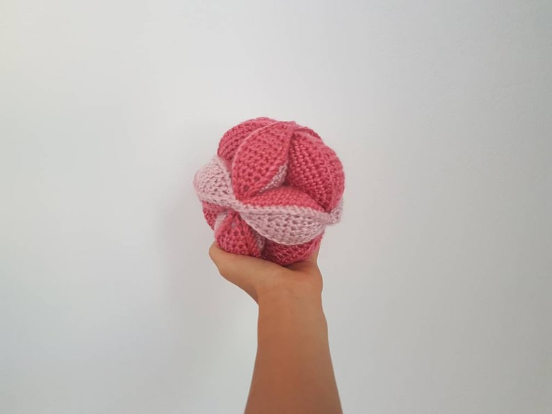 May include: A pink and light pink crocheted ball with a unique, intricate design. The ball is made of yarn and has a soft, textured surface.