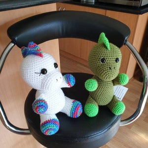 May include: Two crocheted stuffed toys, a white unicorn with rainbow accents and a green dinosaur, sitting on a black swivel chair.