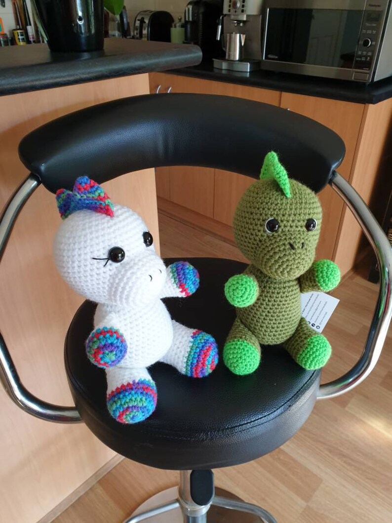 May include: Two crocheted stuffed toys, a white unicorn with rainbow accents and a green dinosaur, sitting on a black swivel chair.