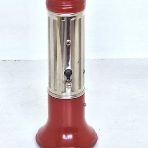 May include: A vintage cylindrical flashlight with a deep red base and top cap, featuring a central silver-toned metal body with vertical ribbed detailing. A small metal switch is positioned on the side. The top cap is embossed with the text Made in Germany.