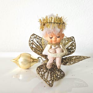 May include: A vintage-style angel figurine with a pale face, closed eyes, and white hair topped with a gold tinsel halo. The figure sits on large, glittery gold mesh wings and is accompanied by a small, smooth, cream-colored teardrop ornament.