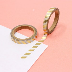 May include: A roll of white and gold washi tape with a diagonal stripe pattern. The tape is on a pink surface and a white surface.