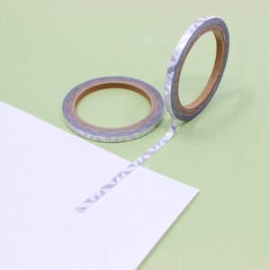 May include: Two rolls of silver washi tape with a geometric pattern. The tape is unrolled and laying on a green surface.