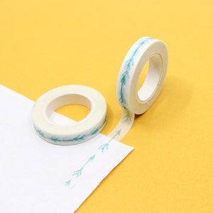 May include: Two rolls of white washi tape with a teal arrow pattern. The tape is unrolled on a yellow surface and a white surface.