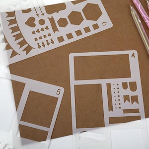Op de afbeelding: Witte plastic sjablonen met diverse vormen en ontwerpen, waaronder harten, pijlen, zeshoeken en rechthoeken. De sjablonen zijn gerangschikt op een bruin oppervlak, met pennen zichtbaar op de achtergrond. Ideaal voor knutselen en journalen.