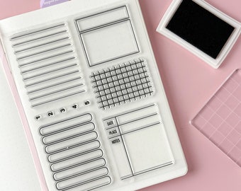May include: Clear acrylic stamps with various designs, including lines, boxes, and a grid, are arranged on a white surface. An ink pad with black ink and a clear acrylic block are also present. The stamps are designed for use in a planner or journal.
