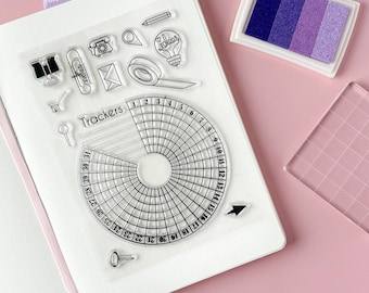 May include: A clear sheet of transparent stamps with various designs, including a lightbulb with the word "ideas" inside, a phone, a tape dispenser, a pencil, a paperclip, a pushpin, an envelope, a magnifying glass, a shopping cart, and a tracker with numbers 1-31. The sheet is on a pink surface.
