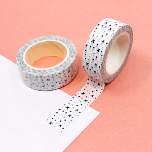 May include: Two rolls of white washi tape with a black and white geometric pattern of dots and crosses. The tape is unrolled and laying on a pink surface.