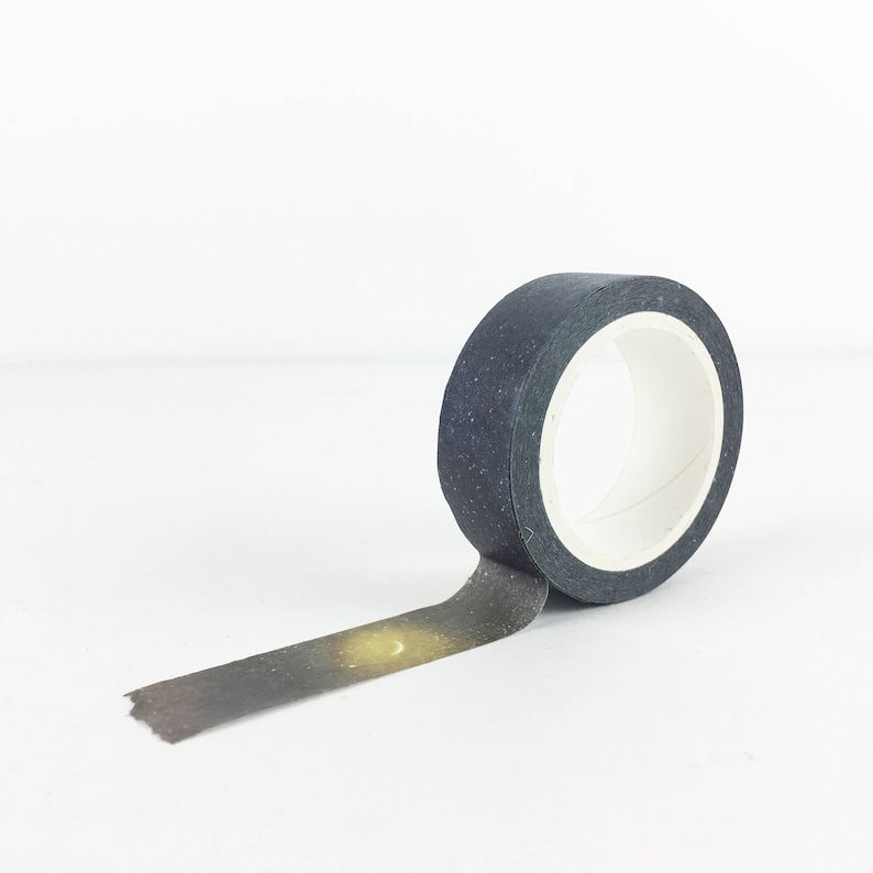 Puede incluir: Un rollo de cinta washi negra con un estampado de galaxia que presenta una luna creciente y estrellas. La cinta est&aacute; parcialmente desenrollada, revelando el dise&ntilde;o.