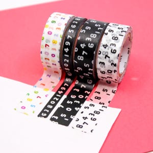May include: Four rolls of decorative washi tape with number patterns. The rolls feature black and white tape with white numbers, and clear tape with colorful numbers. The tape is partially unrolled, revealing the number patterns.