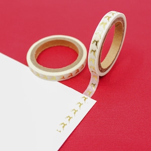 May include: Two rolls of decorative washi tape with a reindeer pattern in gold on a white background. One roll is unraveled, with the tape adhered to a white surface. The background is red.