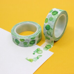 May include: Two rolls of green and white washi tape with a watercolor pattern of green circles. The tape is on a yellow surface and a white surface.
