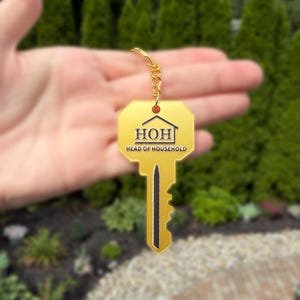 May include: A gold-coloured key-shaped keyring featuring a black outline of a house icon above the text HOH and the words HEAD OF HOUSEHOLD printed in black. The keyring includes a gold metal jump ring and a short gold chain for attachment.