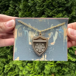 May include: A gold-colored chain necklace with a shield-shaped pendant. The pendant has a layered design with a compass rose and decorative elements. The necklace is displayed on a card with a vintage-style illustration.
