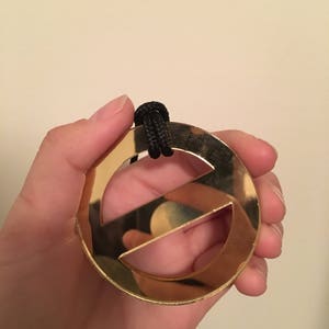 Puede incluir: Un colgante dorado con forma de letra "E" estilizada, suspendido de un cordón negro. El colgante tiene una superficie brillante y reflectante. El diseño es simple y moderno, adecuado para un collar.