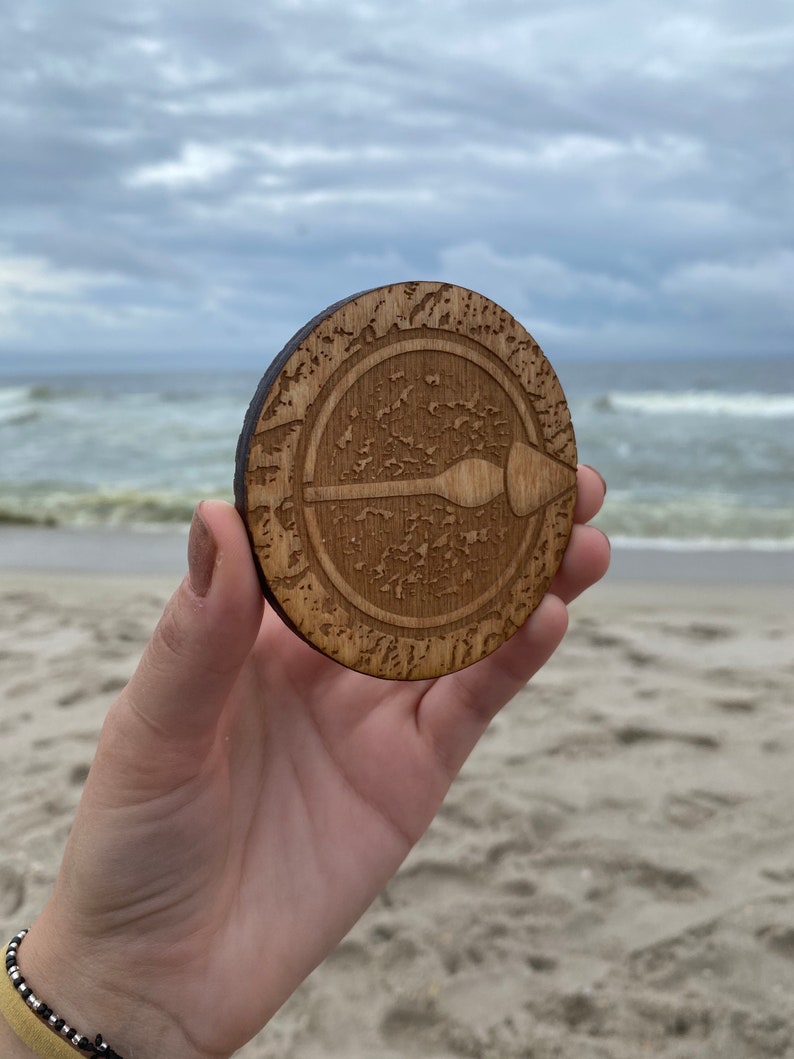 Puede incluir: Un posavasos de madera con un dise&ntilde;o circular que presenta una flecha estilizada que apunta hacia la derecha. El posavasos tiene un acabado de madera de grano marr&oacute;n y est&aacute; rodeado por un c&iacute;rculo de detalles intrincados.