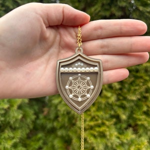 May include: A gold chain necklace with a brown shield-shaped pendant. The pendant has a white star design with white pearl accents.