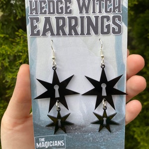 May include: Black acrylic earrings with a star and keyhole design. The earrings are on a white card with the text "Hedge Witch Earrings" and "The Magicians".