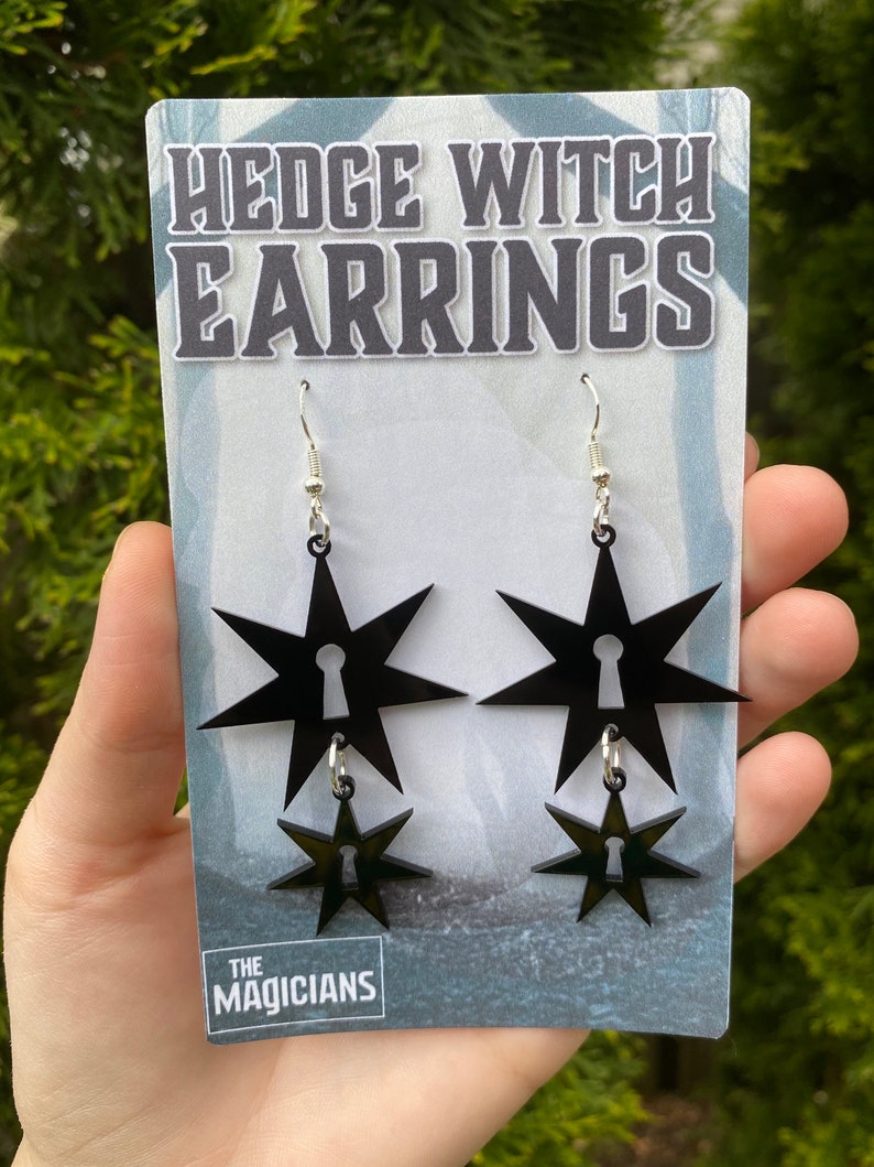 May include: Black acrylic earrings with a star and keyhole design. The earrings are on a white card with the text "Hedge Witch Earrings" and "The Magicians".