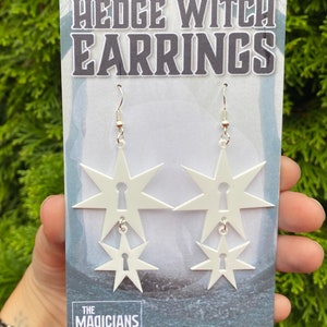 May include: White star-shaped earrings with a keyhole design. The earrings are layered, with a smaller star hanging from a larger star. The earrings are on a white card with the text "HEDGE WITCH EARRINGS" and "THE MAGICIANS".