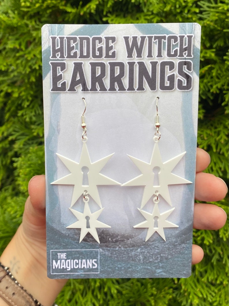 May include: White star-shaped earrings with a keyhole design. The earrings are layered, with a smaller star hanging from a larger star. The earrings are on a white card with the text "HEDGE WITCH EARRINGS" and "THE MAGICIANS".