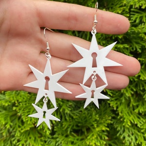 May include: A pair of white star-shaped earrings with a keyhole cutout. Each earring has two stars, one larger than the other.