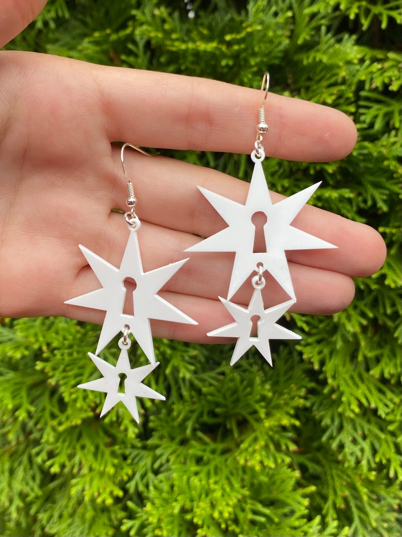 May include: A pair of white star-shaped earrings with a keyhole cutout. Each earring has two stars, one larger than the other.
