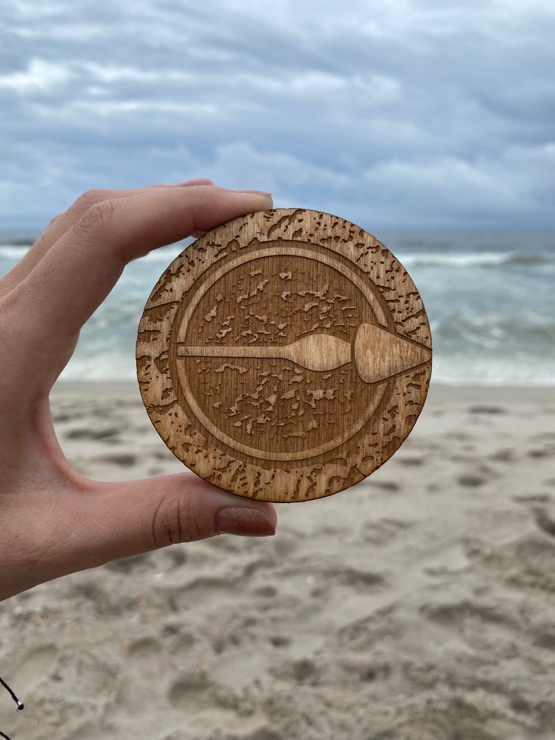 Puede incluir: Un posavasos de madera con un dise&ntilde;o circular que presenta una pala estilizada en el centro. El posavasos tiene un acabado de madera desgastada y est&aacute; rodeado por un borde decorativo.