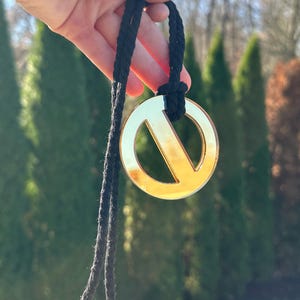 Puede incluir: Un colgante de signo de paz de color dorado en un cordón negro. El colgante es redondo y tiene un diseño de signo de paz recortado.