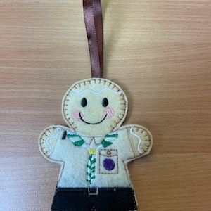 May include: A felt gingerbread person ornament wearing a scout uniform with a green neckerchief and purple badge. The ornament has a brown ribbon for hanging.