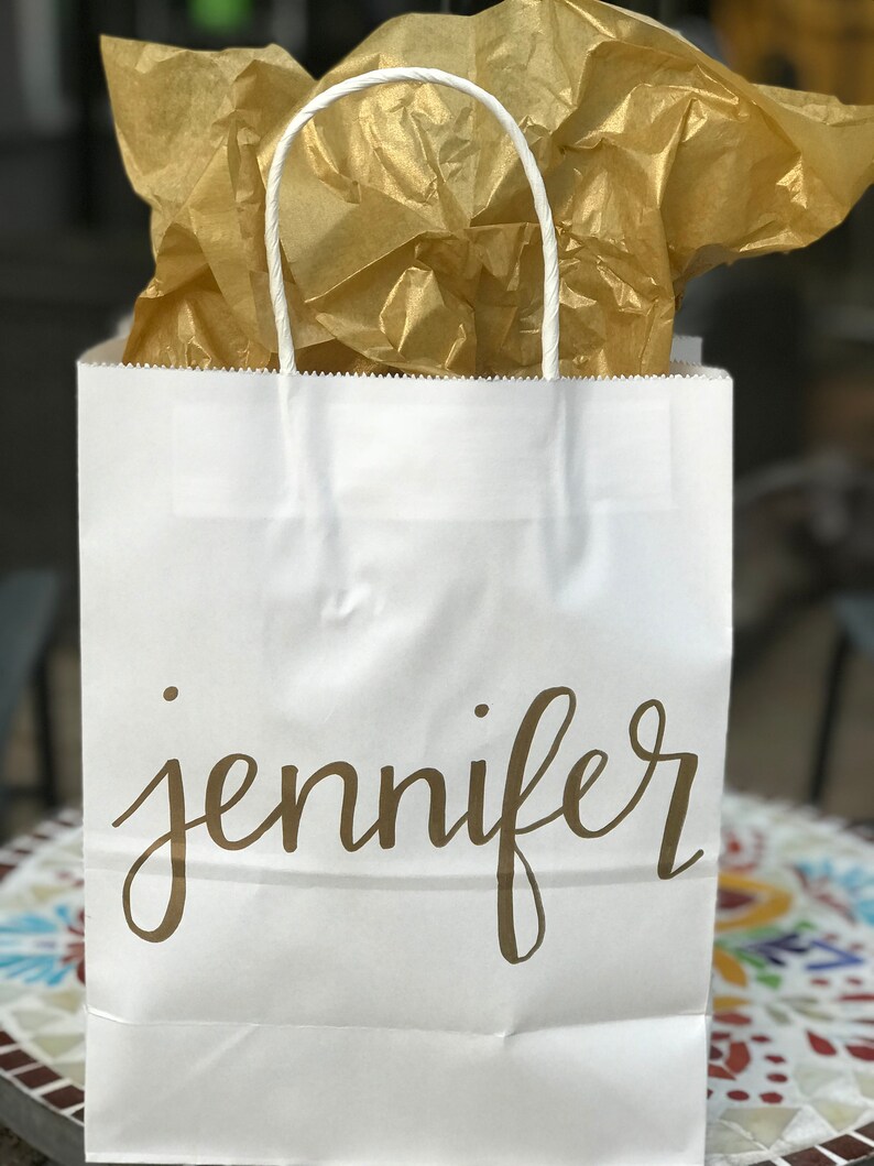 May include: A white paper gift bag with a twisted paper handle. The name "jennifer" is printed in gold script on the front. The bag is filled with gold tissue paper, suggesting a celebratory or gift-giving occasion.