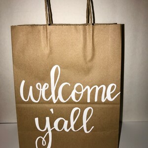 May include: A brown paper gift bag with twisted paper handles. The words "welcome y'all" are written in white script across the front of the bag. The bag is empty and ready to be filled.