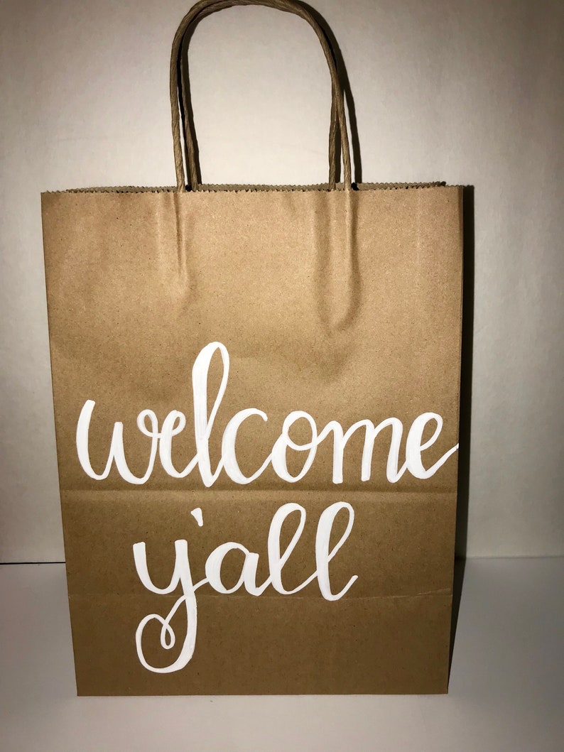 May include: A brown paper gift bag with twisted paper handles. The words "welcome y'all" are written in white script across the front of the bag. The bag is empty and ready to be filled.