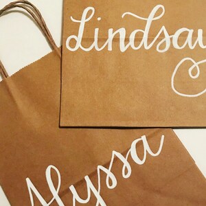 May include: Two brown paper gift bags with white cursive lettering. One bag reads "Lindsay" and the other reads "Alyssa". The bag on the left has a twisted brown paper handle.
