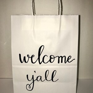 May include: White paper gift bag with twisted paper handles. The bag features the words "welcome y'all" in black cursive script. The bag is designed for gifting and is suitable for various occasions.