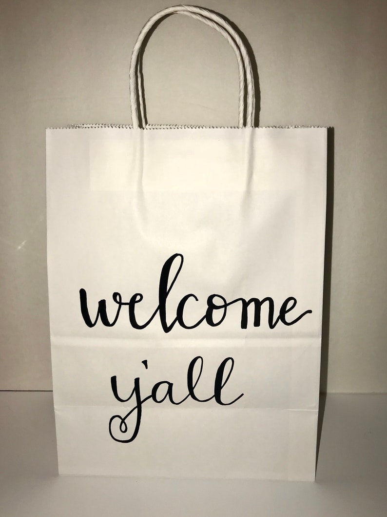 May include: White paper gift bag with twisted paper handles. The bag features the words "welcome y'all" in black cursive script. The bag is designed for gifting and is suitable for various occasions.