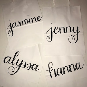 May include: Four white paper gift bags with twisted paper handles. Each bag has a name in black cursive script: "jasmine", "jenny", "alyssa", and "hanna". The bags are arranged on a light-colored surface.