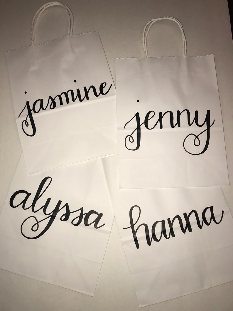 May include: Four white paper gift bags with twisted paper handles. Each bag has a name in black cursive script: "jasmine", "jenny", "alyssa", and "hanna". The bags are arranged on a light-colored surface.